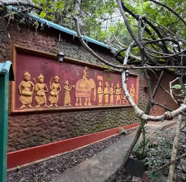 A carved frieze of a procession with an elephant, on a wall among the trees